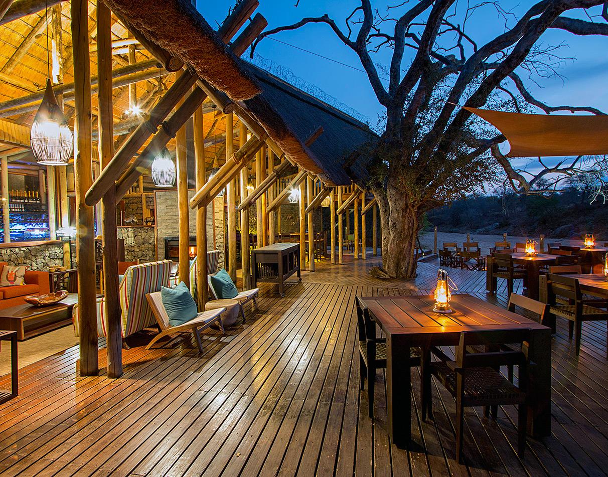 Rhino Post Safari Lodge open deck at dusk with lantern-lit dining tables, wooden beams and riverbed views.