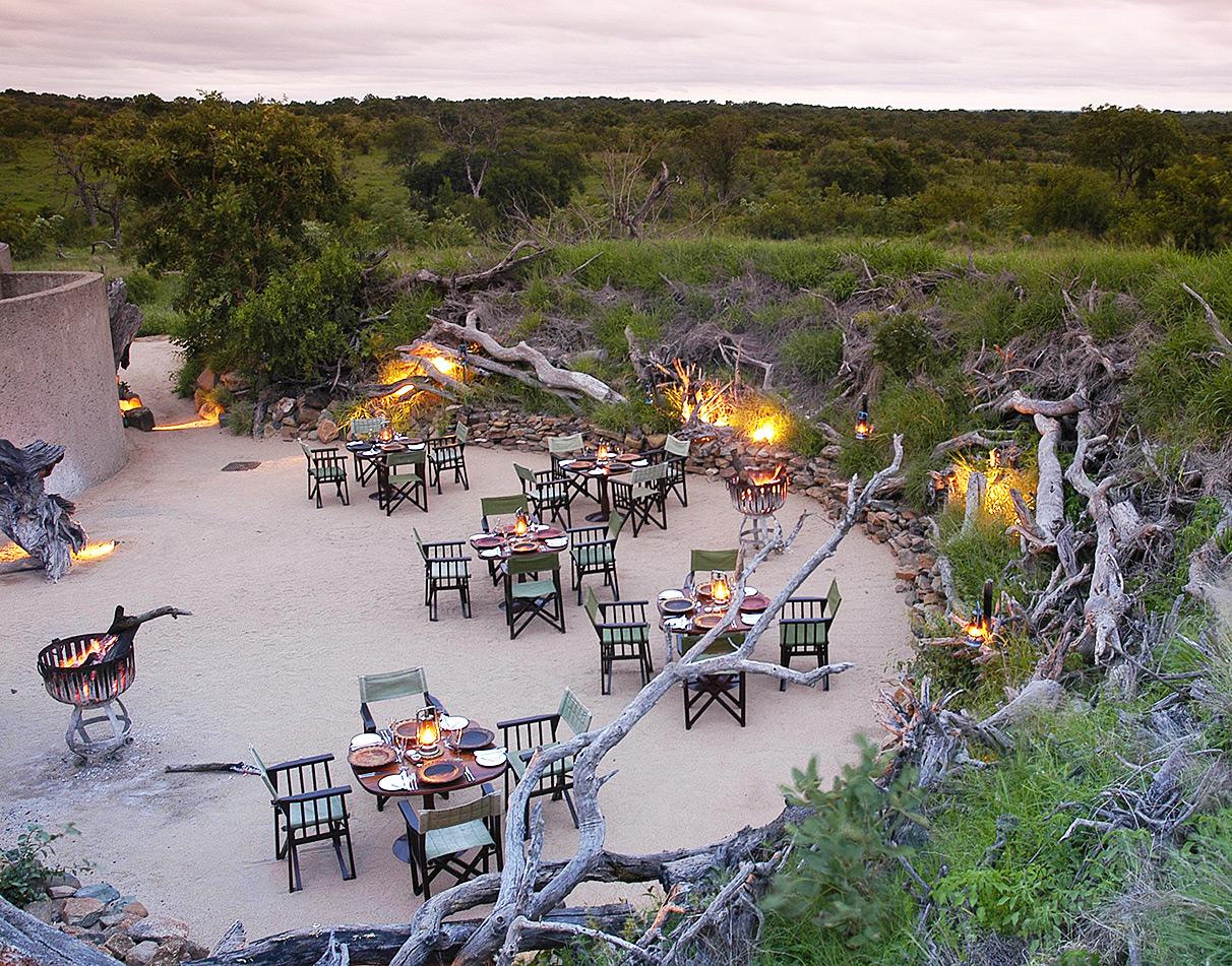 Sabi Sabi Earth Lodge outdoor bush dining experience at dusk with lanterns, fire baskets and tables set in a dry riverbed.