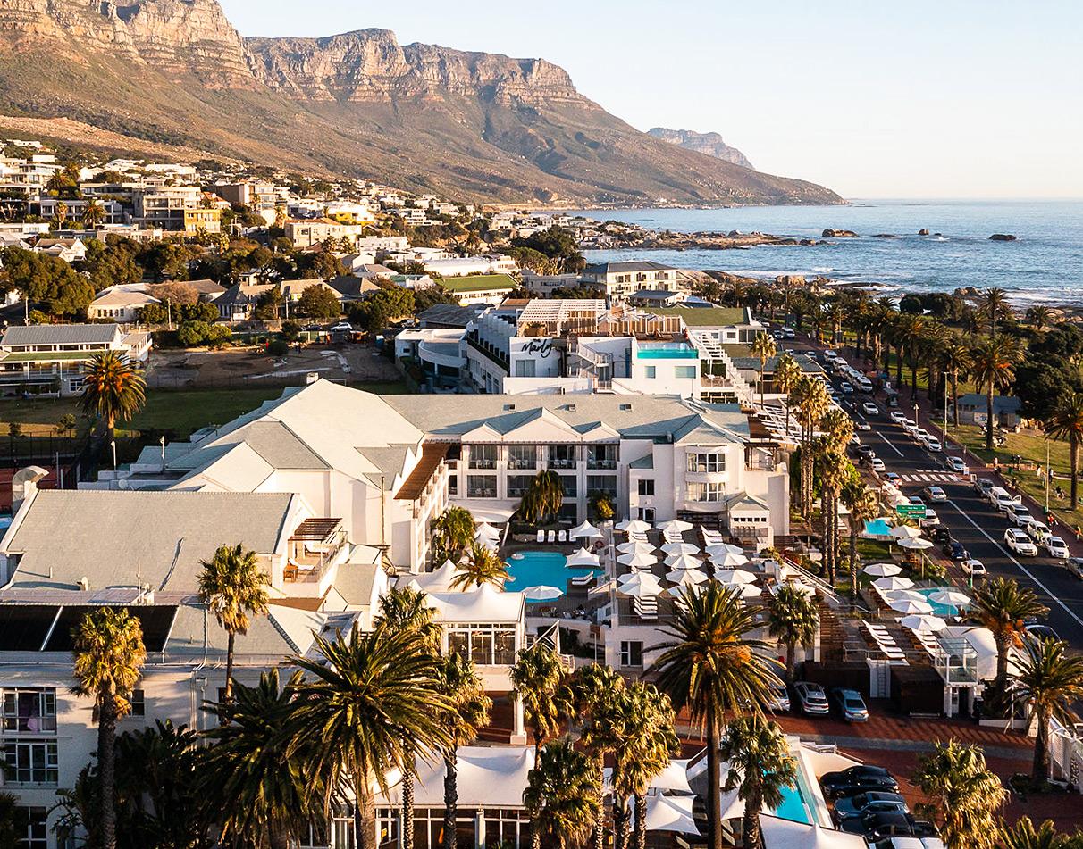 Aerial view of The Bay Hotel with pool and palm trees beneath the Twelve Apostles mountain range, Camps Bay, Cape Town.