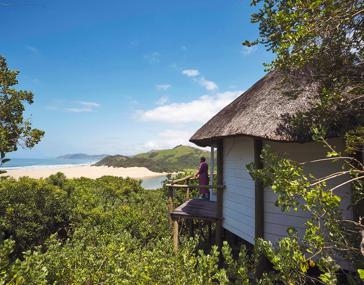 Umngazi River Bungalows thatched cottage nestled in the forest with a guest enjoying panoramic views over the estuary and beach.