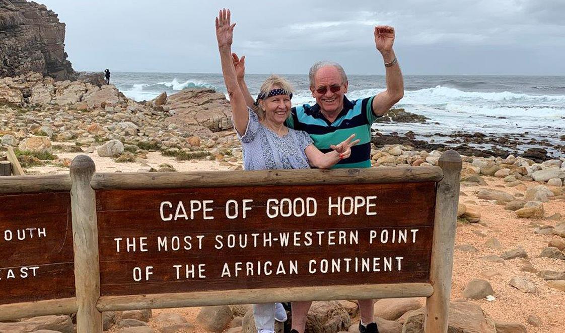 A couple stand behind the Cape of Good Hope sign, smiling with their arms int he air.