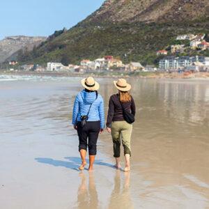 Two women walking barefoot in shallow water along the beach of Muizenberg near Cape Town.