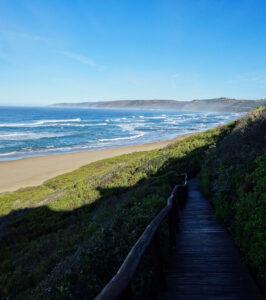 Wooden boardwalk leading down to a large sandy beach with ocean waves on the Garden Route, South Africa.