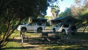 Two guided self-drive vehicle stand at a campsite with tables and chairs set up.