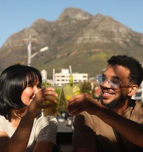 Young couple cheersing their mojitos at a bar in Cape Town with stunning views of the mountains in the background.