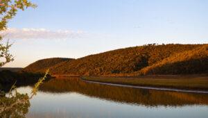 Golden hour light over a tranquil lagoon surrounded by forested hills along the Garden Route, South Africa.