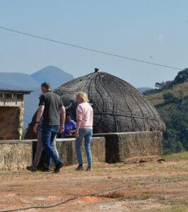 Couple visiting a traditional Swazi village with a thatched hut and mountain backdrop in Eswatini, ensure you include gratuities for the staff in your South Africa travel budget.