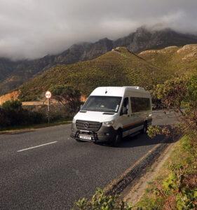 A small group tour vehicle driving up a hill with mountains and clouds in the background.