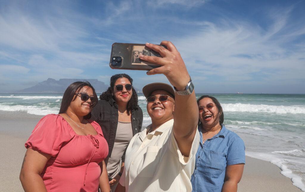 A group of four girls smile at the camera as they take a selfie at the beach with Table Mountain in the background.