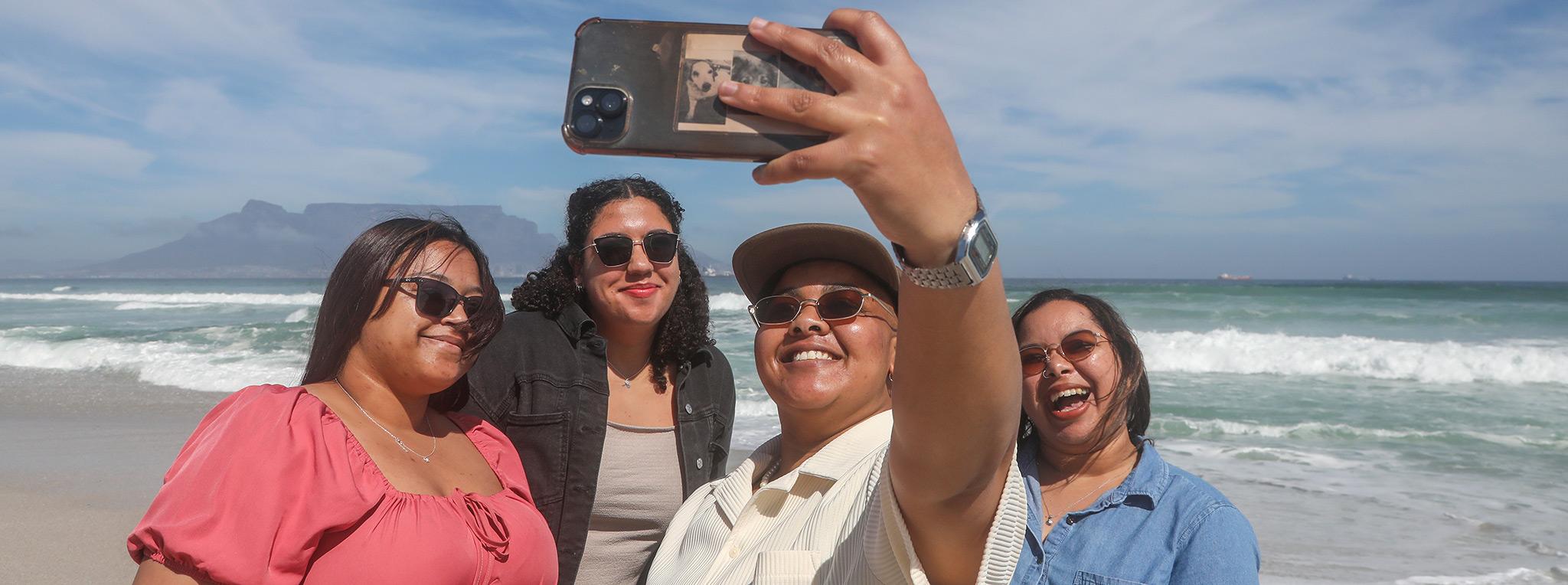 A group of four girls smile at the camera as they take a selfie at the beach with Table Mountain in the background.