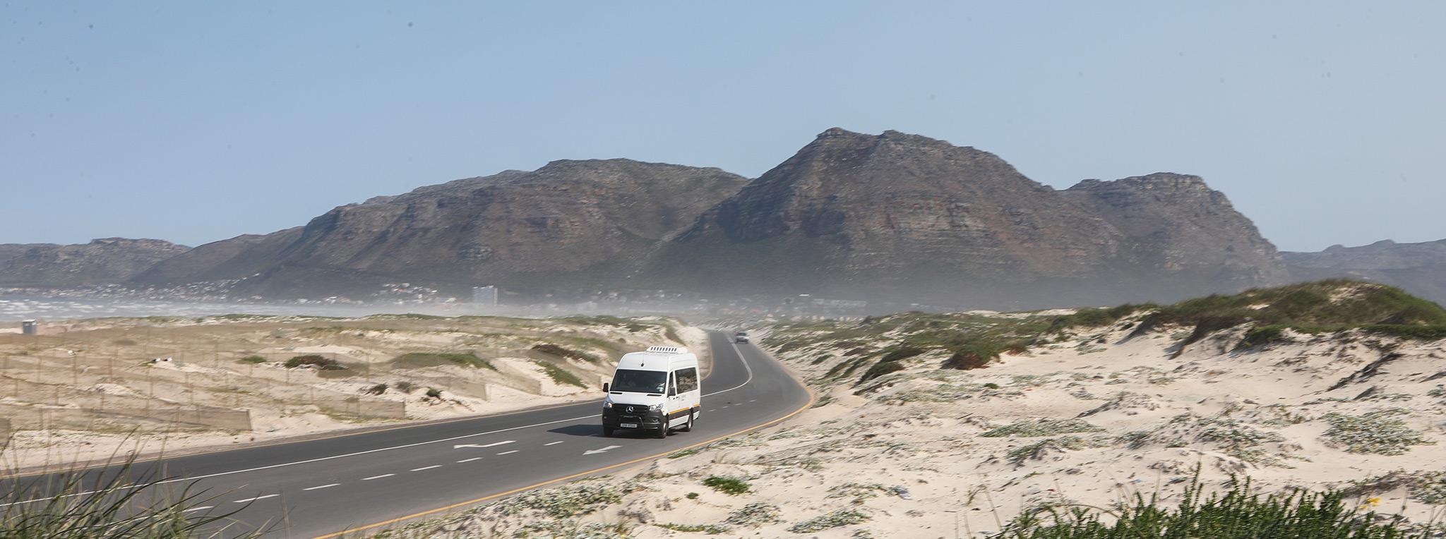 A Mercedes Sprinter tour vehicle driving along a coastal road through sand dunes and mountains, South Africa.