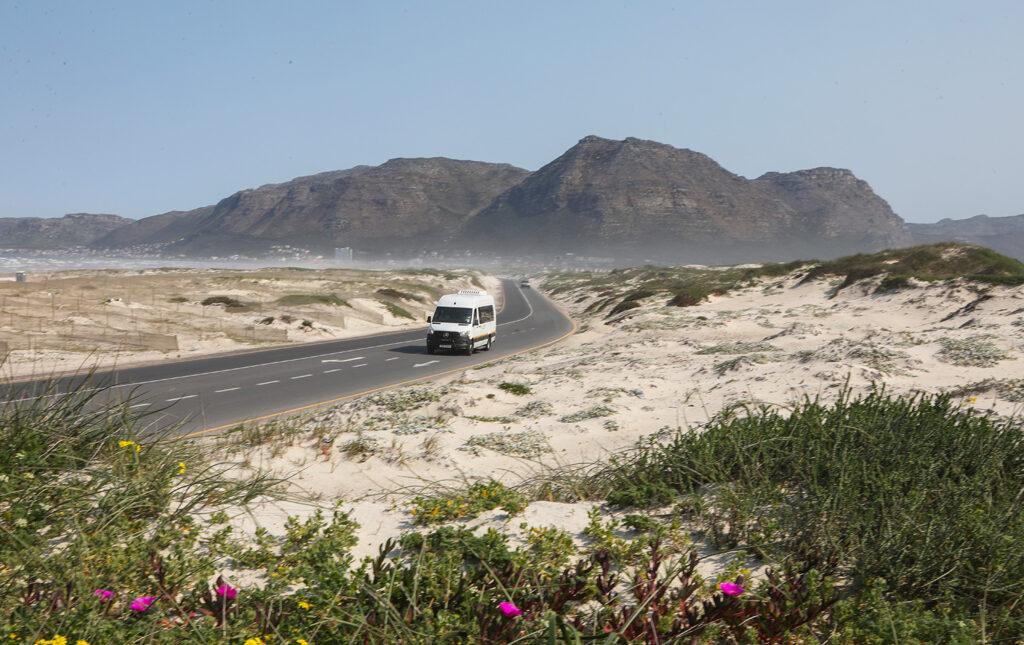 A Mercedes Sprinter tour vehicle driving along a coastal road through sand dunes and mountains, South Africa.