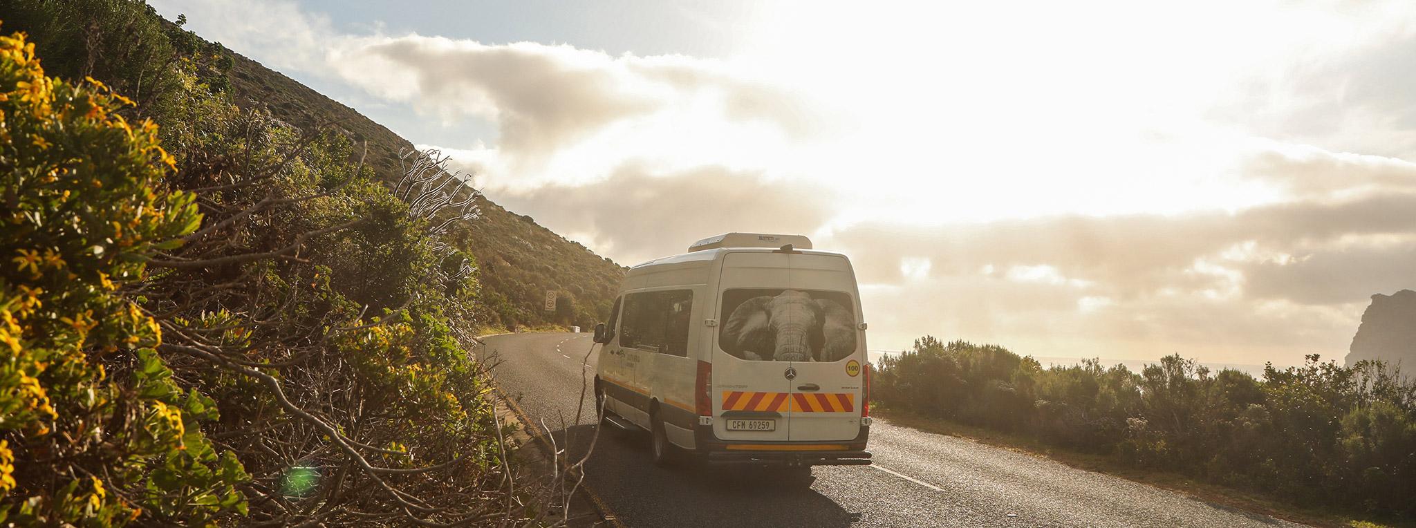 A tour vehicle on a guided South Africa tour drives along a coastal road at sunset.