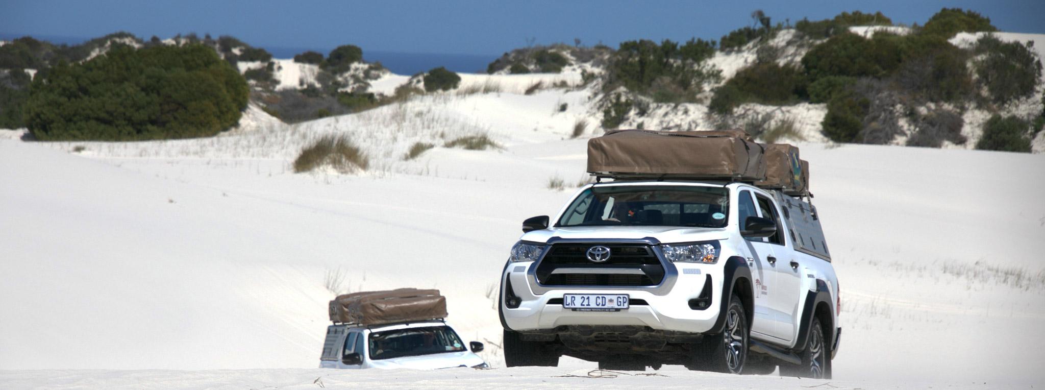 Two white 4x4 vehicles with rooftop tents drive through Atlantis dunes on a guided self-drive tour in South Africa.