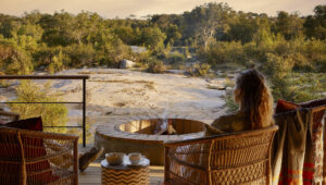 Woman having a cup of coffee sitting on a wooden deck with fire pit overlooking the Kruger Park bush.