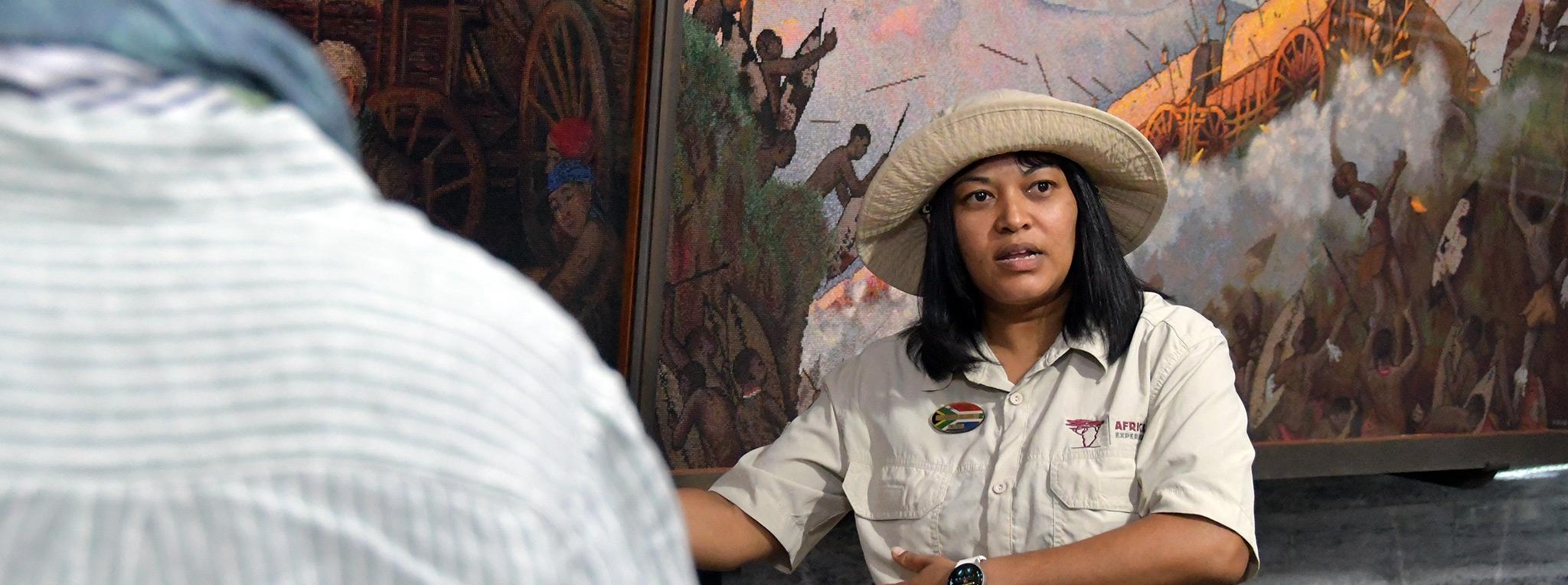 Tour guide explaining the history of the Great Trek at the Voortrekker Monument in Pretoria, South Africa, with historic paintings in the background.