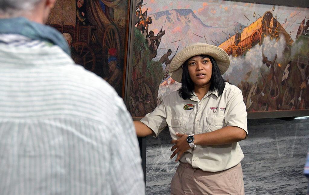 Tour guide explaining the history of the Great Trek at the Voortrekker Monument in Pretoria, South Africa, with historic paintings in the background.
