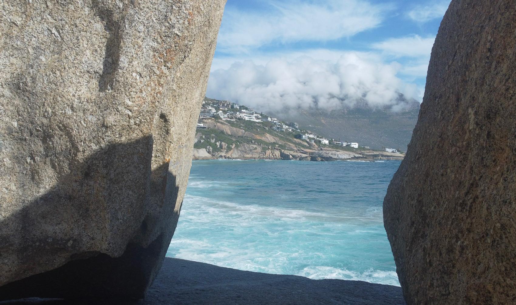 Coastal view of Cape Town with houses on a hill, framed by large rocks.