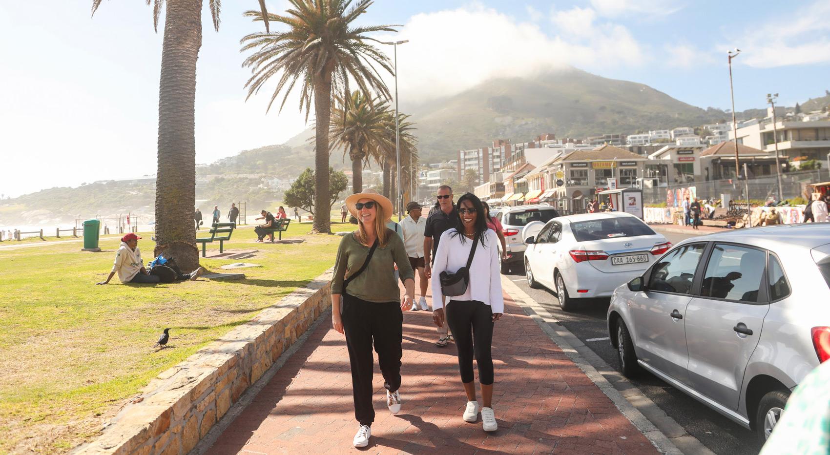 People walk along a sunny promenade in Cape Town, with palm trees and mountains in the background. People walk along a sunny promenade in Cape Town, with palm trees and mountains in the background.